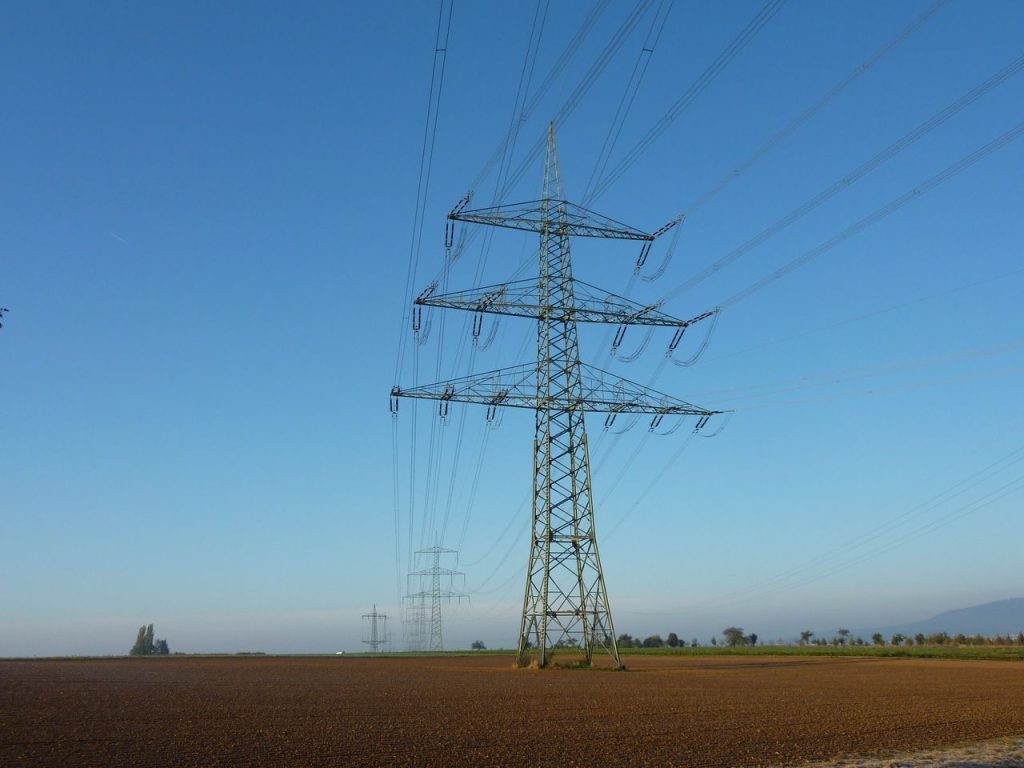 High voltage power lines on a tower standing in an open field against a clear blue sky.